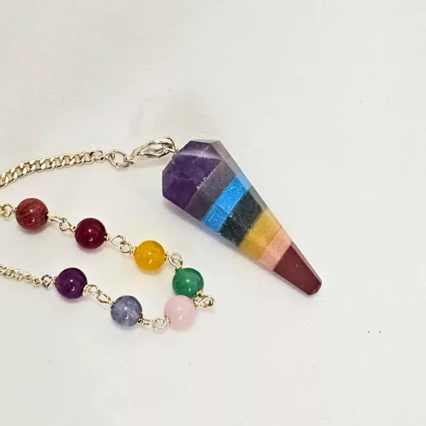 Vavatu 7 Chakra Crystal Pendulum is shown. The colors are purple, grey, blue, yellow, beige, red, and black, and it is a 6 faceted diamond shaped. The crystal pendulum is in the center of the photo with a white backdrop and a single thin gold chain attached to the larger circular end with a small bead of each of the stones included in the pendulum incorporated into the chain.