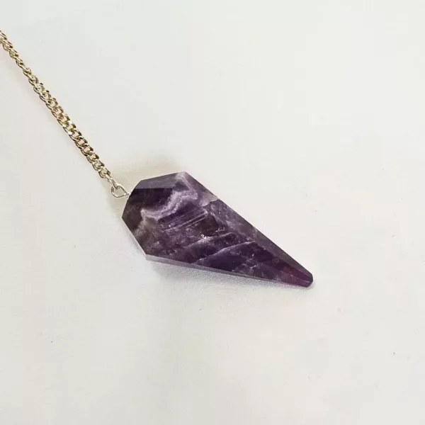 Vavatu Amethyst Crystal Pendulum is shown. The color is purple, and it is a 6 faceted diamond shaped. The crystal pendulum is in the center of the photo with a white backdrop and a single thin gold chain attached to the larger circular end.